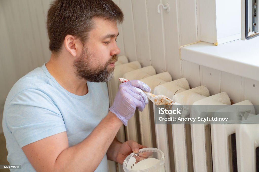 man in glove painting heating radiator in home man in a glove painting a heating radiator with a paint roller in home. A DIY repair. Professional Workman Hand holding Dirty Paintbrush. Painting - Activity Stock Photo