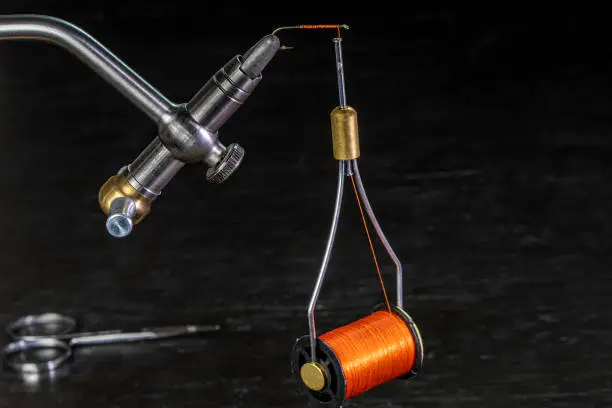 bobbin of thread hanging from a hook on a fly tying vise and a pair of scissors on the table