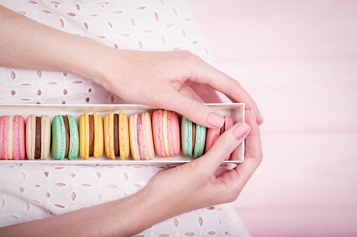 Beautiful female hands holding gift box of delicious french colorful macaroons on pale pink background. Top view, copy space