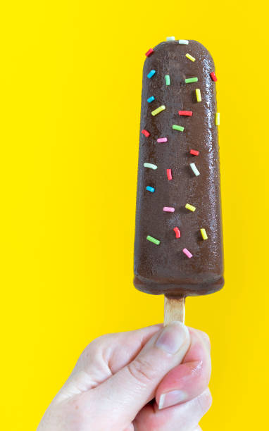 mano della donna che prende un gelato al cioccolato ricoperto di pepite colorate su sfondo giallo - studio shot gourmet freshness cold foto e immagini stock