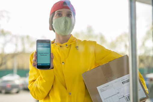Young female courier in workwear and mask showing transaction data on smartphone screen while waiting for client by the door