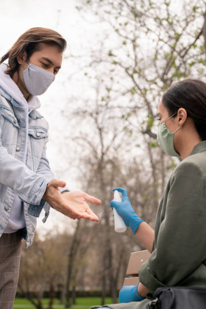 istock Young man in mask opening palms while his girlfriend spraying sanitizer 1225197368