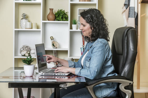 Adult woman using laptop with pleasure in light contemporary apartment Side view of joyful lady in denim clothes focusing and point finger on screen while sitting at table in soft leather armchair and using laptop at home home office chair stock pictures, royalty-free photos & images