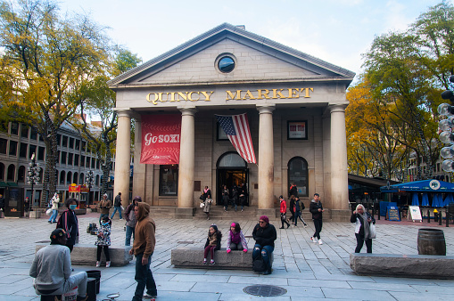 Boston, Massachusetts.  October 30, 2018.  People ouside Quincy market with a red sox banner and american flag hanging between the buildings pillars on an autumn day.