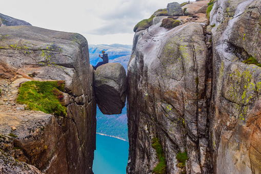The tourist hiker man take a selfie while sitting on top of Kjeragbolten - the most dangerous stone in the world. Kjeragbolten is a rock stuck at an altitude of 984 meters above Lysefjorden, Norway.