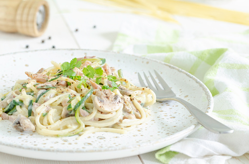 focus on a single serving plate of carbonara style with spaghetti, zucchini,mushroom,bacon and cream on a white wooden table with peeper shaker and pasta on background