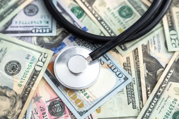 Photo of Doctor or nurse stethoscope medical device or equipment with metal parts and black tubing laying on a pile of united states currency in large bills or cash covering the entire surface.