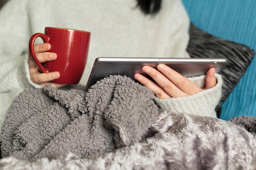 Woman on sofa covered with blanket looking at tablet and holding hot drink mug