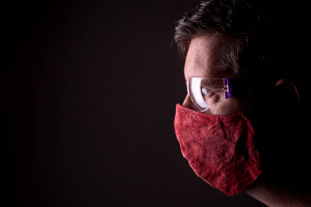 istock Profile of a caucasian male looking straight forward through reflecting protective plastic glasses and a homemade wrinkled red cloth face mask on. 1225235499