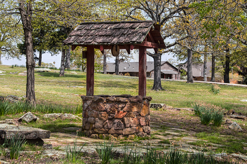 Rustic rock and wooden wishing well surrounded by paving stones and trees in springtime with log cabin in distance