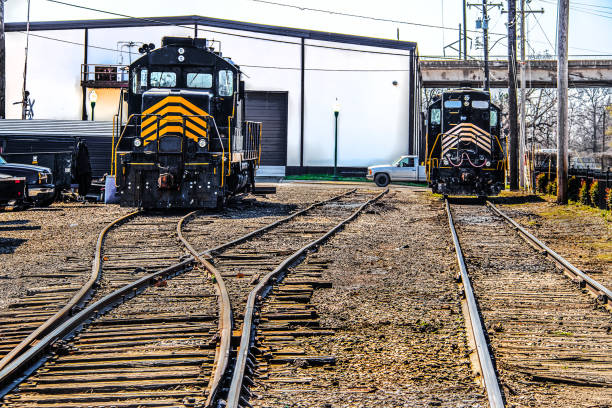 two train engines sitting in a train yard with big white shop behind them and many tracks crisscrossing in foreground - shallow focus - connection merger road togetherness imagens e fotografias de stock