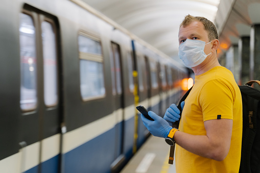 Male worker uses modern smartphone on platform, waits metro or train, wears surgical mask for virus protection, keeps social distancing to crowd, cares about health, commutes by public transport
