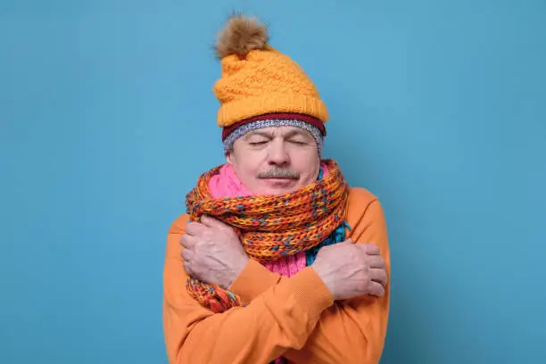 Upset senior hispanic man wearing several hats and scarfs freezing because of cold weather. Studio shot on blue wall.