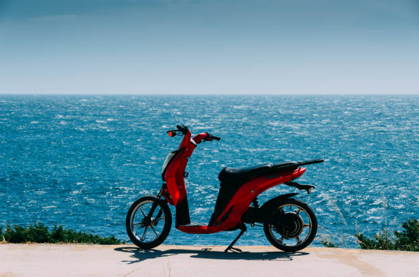 red scooter parked on a green grassy beach next to the ocean on a sunny summer day with a blue sky - vespa scooter imagens e fotografias de stock