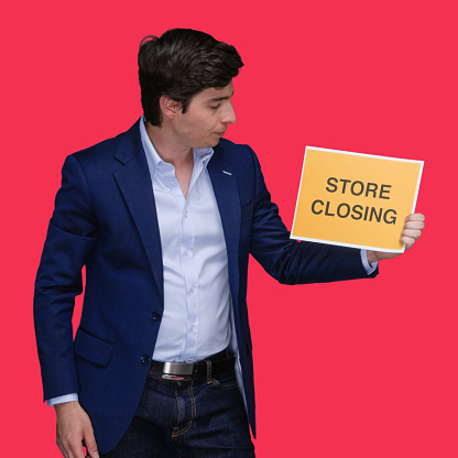 Waist up of aged 20-29 years old with short hair caucasian young male standing in front of colored background in the store wearing shirt who is bankrupt and holding banner sign