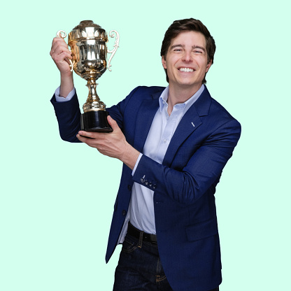 Front view of aged 20-29 years old with short hair caucasian young male standing in front of colored background wearing jacket who is successful and winning and showing award who is in first place and holding trophy