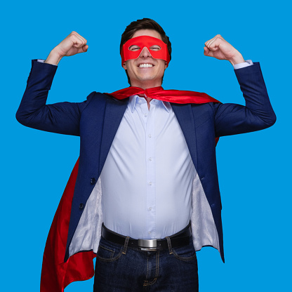 Waist up of with short hair caucasian young male superhero standing in front of blue background wearing cape - garment who is conquering adversity and showing flexing muscles