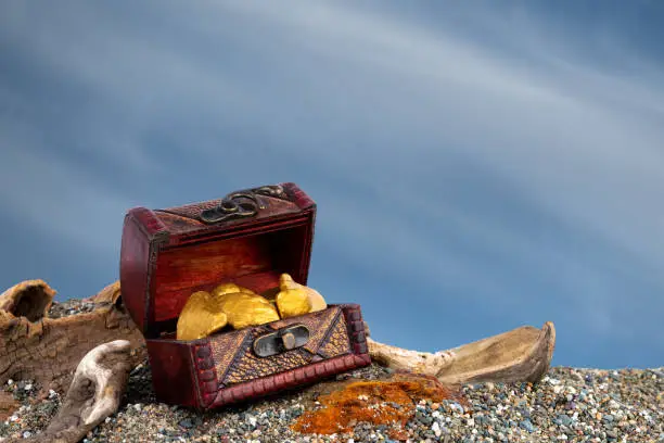 A small treasure chest filled with gold nuggets. Chest is on a sandy beach with driftwood. Background is blue sky with clouds.