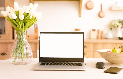 Front view of laptop with white screen in a kitchen with soft and warm light.help in the kitchen with new technologies.isolation at home by the coronavirus concept . freelance. flowers on the table.