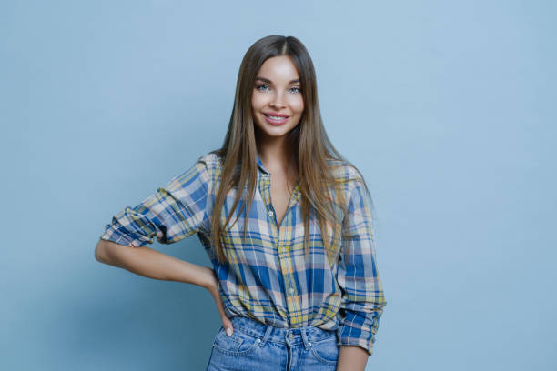 istock Charming pretty millennial lady keeps hand on figure, stands confident and smiles cheerfully, wears checkered shirt and jeans stands in casual posture happy to meet with friend enjoys pleasant talk 1225264910