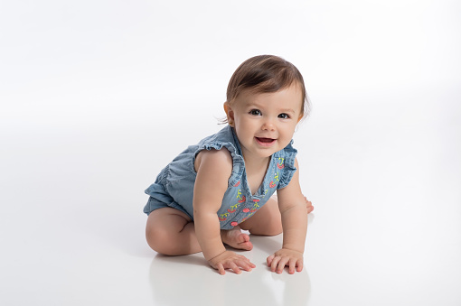 A smiling six month old baby girl wearing a denim romper. She is sitting on a white, seamless background and attempting to crawl.
