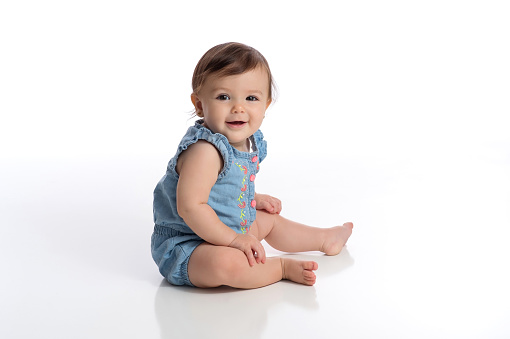 A smiling eight month old baby girl wearing a denim romper. She is sitting on a white, seamless background and looking at the camera.