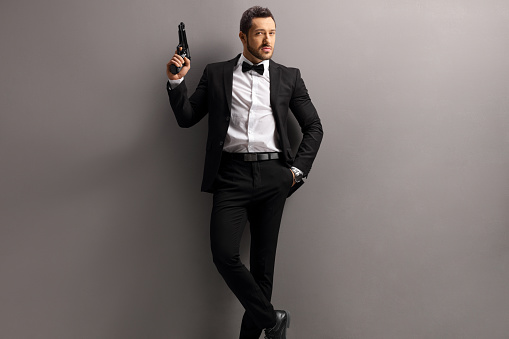 Man in a suit holding a gun and leaning on a gray wall