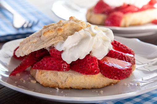 A serving of strawberry shortcake with whipped cream on a white plate. There is another serving of plated shortcake in the background. The depth of field is shallow, and focus is on the berries in the front.