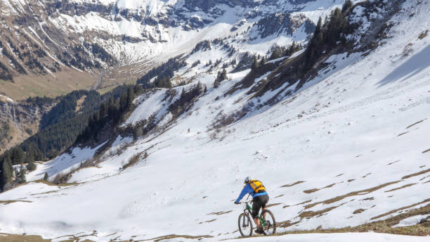 istock Mountain biker rides down snowy mountain ridge 1225280022