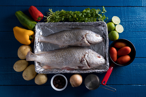 Preparing Brazilian Corvina Fish with Red Peppers, Green Pepper, Yellow Pepper, Parsley, Potato, Black Olive, Onion, Italian Tomatoes and Lemon on the Marine Blue Wood Table