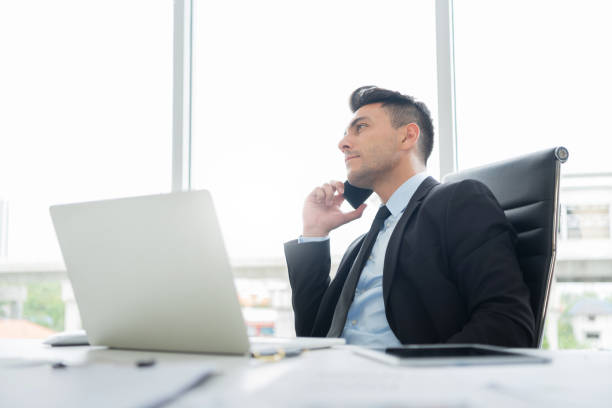 istock Smart Handsome Businessman in black suit is sitting using a mobile phone for negotiating business deals with partners and clients while working on a laptop computer at the workplace in the office. 1225319840
