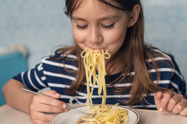 彼女の顔に満足した表情をした子供の女の子は、自宅の台所で夕食を食べます。スパゲッティを食べるかわいい面白い小さな女の子 - child eating pasta spaghetti ストックフォトと画像