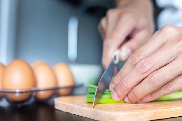 closeup of people hand and knife slice spring onion on wood butcher and fresh chicken egg in bowl and group of egg in box on wooden kitchen table for healthy and delicious meal or food cook concept for family dinner menu