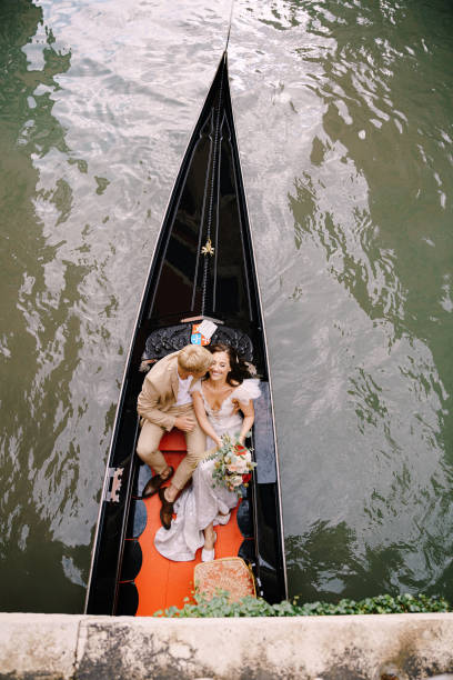 italien hochzeit in venedig. ein gondeltier rollt braut und bräutigam in einer klassischen holzgondel entlang eines schmalen venezianischen kanals. brautpaare sitzen in einem boot auf dem hintergrund alter gebäude. - shoe groom wood luxury stock-fotos und bilder