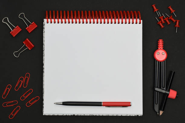 Red stationery items and blank spiral notebook on a black background. Stationery set. Geometric drawing compass, paper clips, stationery pin around white paper notebook with pen.