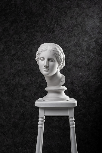 Gypsum copy of ancient white statue of bust of Venus on chair with textured black background .Plaster sculpture woman face. The goddess of love in Greek mythology. Renaissance epoch