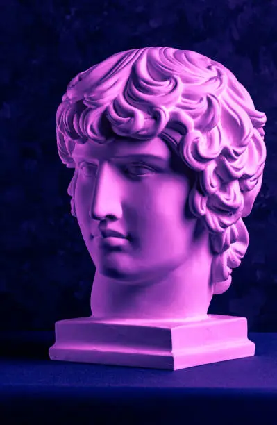 Photo of Gypsum copy of ancient famous statue Antinous head on dark textured background. Plaster antique sculpture young man face Renaissance epoch. Purple toned.