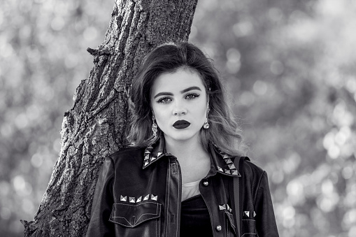 Black and white portrait of beautiful stylish young woman, wearing black leather jacket, standing in a park near the tree.