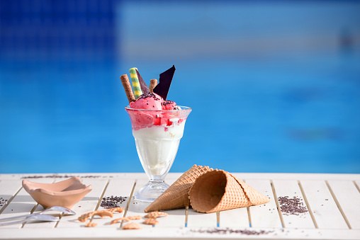 Panorama of glass with vanilla, watermelon ice cream on blur bokeh background with copy space. Cherry berries sorbet with waffle cones and chocolate piece on white table top of blue water pool