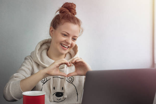 молодая кавказская женщина с ноутбуком дома делает видеозвонок, разговаривает и улыбается, показывая жест формы сердца. оставайтесь на свя - having ones say стоковые фото и изображения