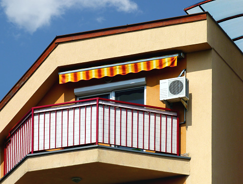 Low angle view of balcony with canvas sunshade canopy in yellow and red. steel picket guard rail. blue sky and white clouds. air conditioner condenser unit mounted on the wall. glass canopy on the side. home ownership concept.