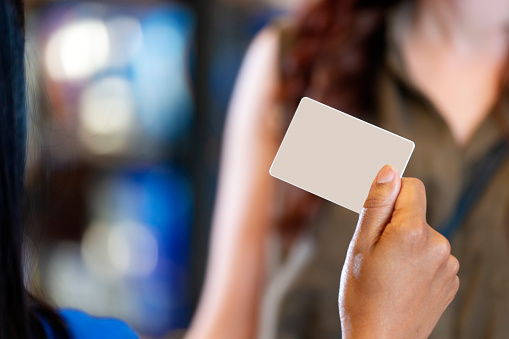 A female hand holds a blank ID card or credit card in front of a retail checkout till computer screen with an unrecognizable person in the background.