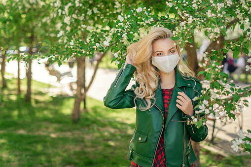 Blonde girl in a white medical mask and a green jacket straightens her hair, standing under a blossoming apple tree