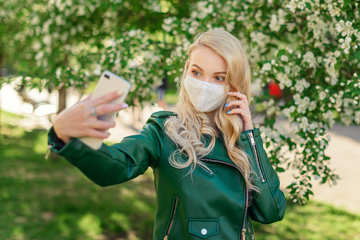 A blonde girl in a white medical mask and a green jacket holds a white smartphone in her hands and takes a selfie, standing next to a blossoming apple tree