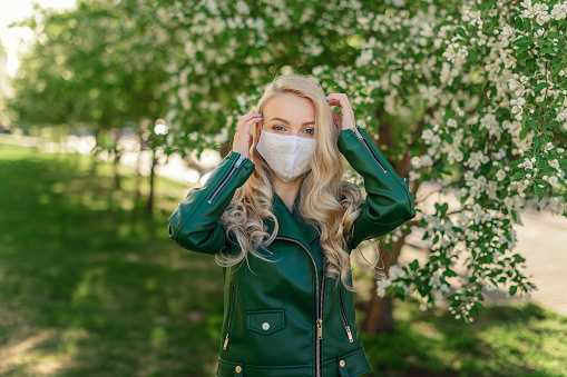 Blond girl in a white medical mask and a green jacket straightens her hair, standing near a blossoming apple tree