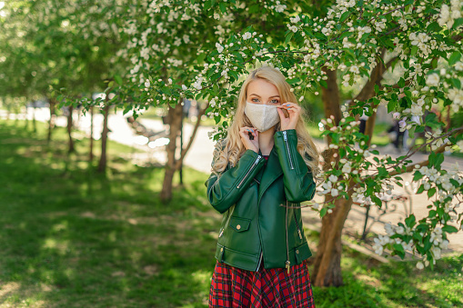 A blonde girl with beautiful eyes stands in the open air near blooming apple trees and adjusts the protective mask against coronavirus on her face.