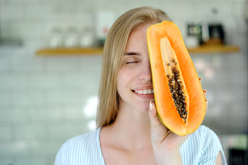Concept of summer and seasonal food. Portrait view of carefree young adult woman covered half face with papaya fruit, smiling wide and standing on kitchen with copy space
