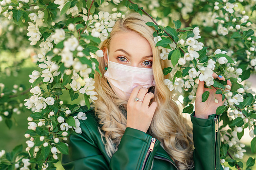 Blond girl in a white medical mask and a green jacket adjusts the mask on her face with her hand, holding the branches of a blossoming apple tree with her other hand