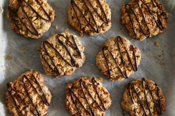 Image of baked homemade carrot, coconut and chocolate chip cookies drizzled with dark chocolate, on parchment paper / greaseproof paper on non-stick baking oven tray, elevated view Stock photo showing an elevated view of greaseproof paper lined baking oven tray containing freshly baked, homemade carrot, coconut and chocolate chip biscuits, home baking concept. chocolate chip cookie top view stock pictures, royalty-free photos & images