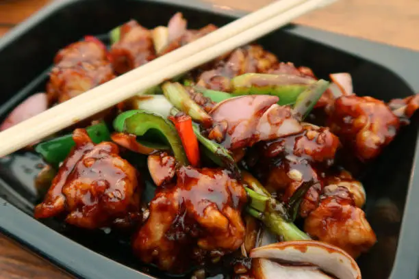 Photo of Image of spicy Kung Pao Chicken takeaway meal in black plastic disposable container with wooden chopsticks, onion, red and green peppers, elevated view